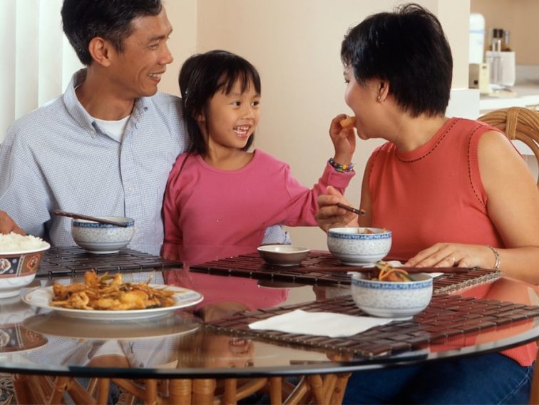 An Asian family, an adult male and female are seated around a table eating a meal with a young female standing in between the adults. Photographer Rhoda Baer